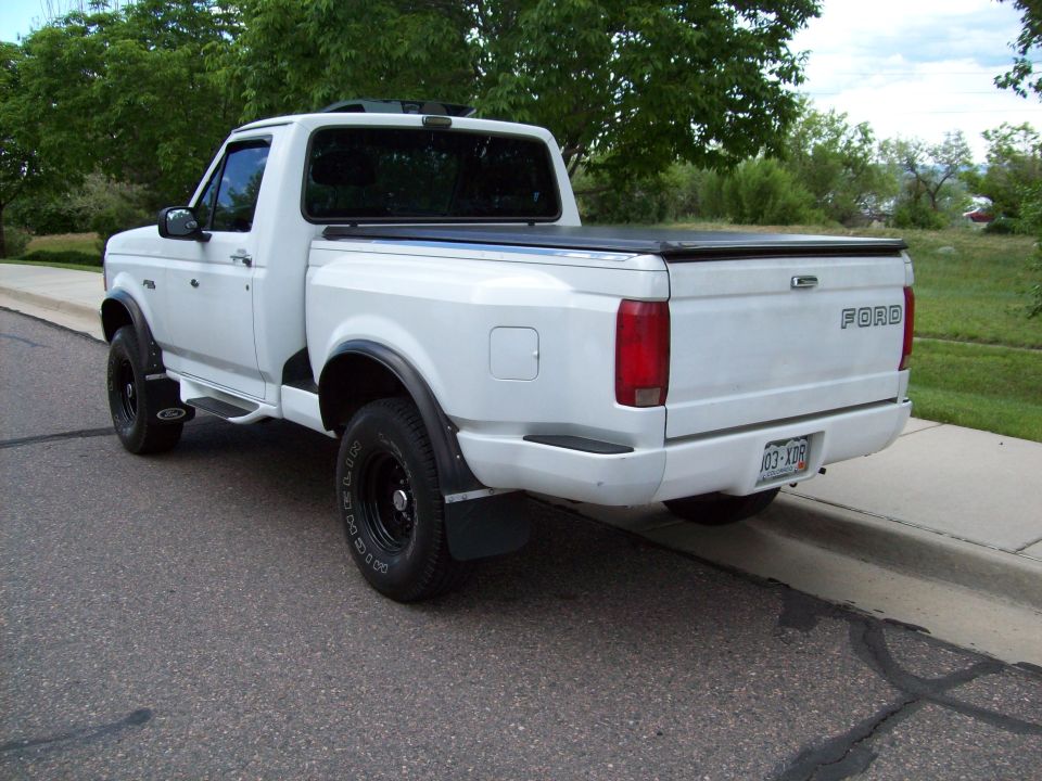 White Ford F-150 pickup truck, rear 3/4 view, parked on asphalt street, black tonneau cover, aftermarket wheels