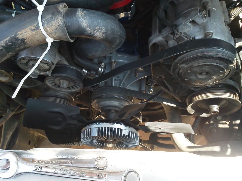 Engine bay showing fan clutch, belts, pulleys, and hoses. Two wrenches are visible in the foreground