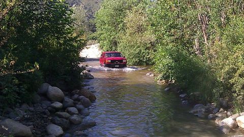 1988 Ford Bronco - A red SUV navigates a rocky riverbed surrounded by lush green foliage.