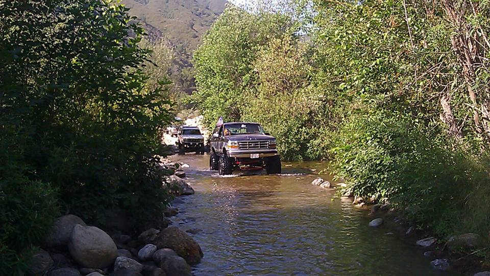 Dark gray 1992 Ford Bronco driving through a shallow river, lifted suspension, large tires, trees and rocks line the banks