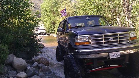 1988 Ford Bronco - A lifted dark blue SUV navigates a rocky stream, with another vehicle visible in the background.