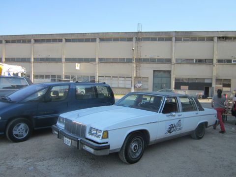 1991 Ford Crown Victoria - A white sedan with a dark roof is parked next to a blue minivan in front of a large industrial building.