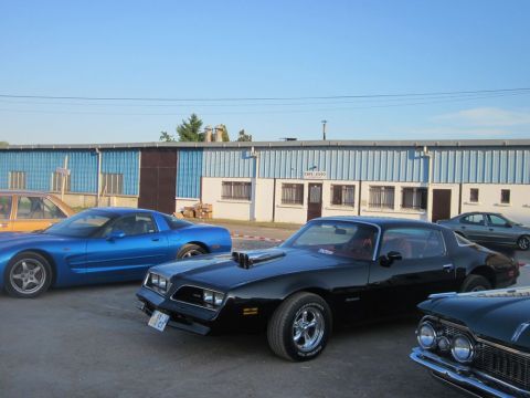 1991 Ford Crown Victoria - A black coupe with a prominent hood scoop is parked alongside a blue Corvette and other vehicles in front of a building.