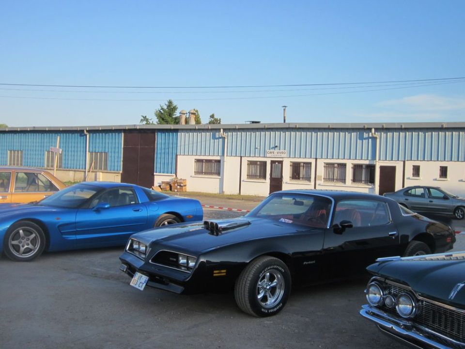 Black coupe with hood scoop, chrome wheels, and custom exhaust pipes. Blue Corvette and other cars visible