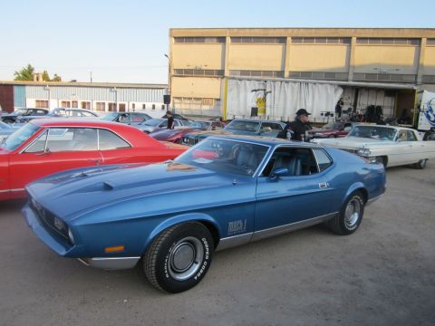 1991 Ford Crown Victoria - A blue 1971 Ford Mustang Mach 1 is parked among other vehicles, with a building and truck in the background.