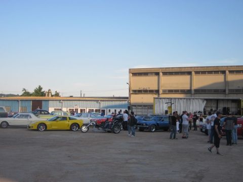 1991 Ford Crown Victoria - A gathering of vehicles and people in an outdoor lot with a large industrial building behind them.