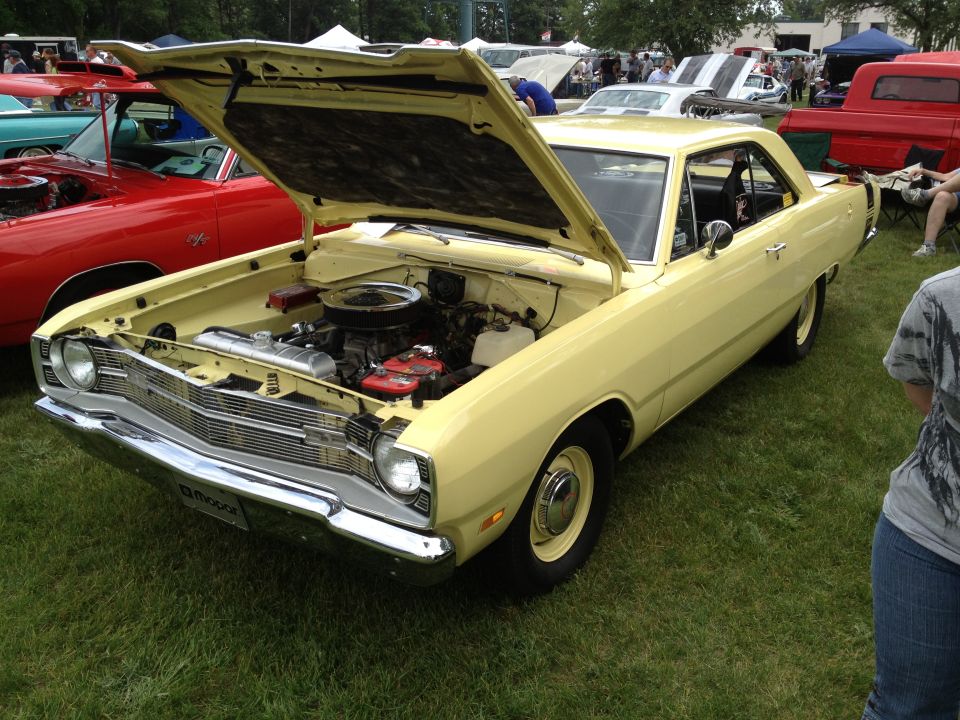 Yellow coupe with hood open, showing engine and front grille. A red car is parked to the left