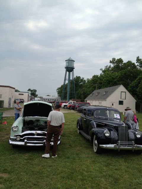 1988 Ford Bronco - A car show features a light green sedan and a dark sedan under a cloudy sky, with a water tower visible.