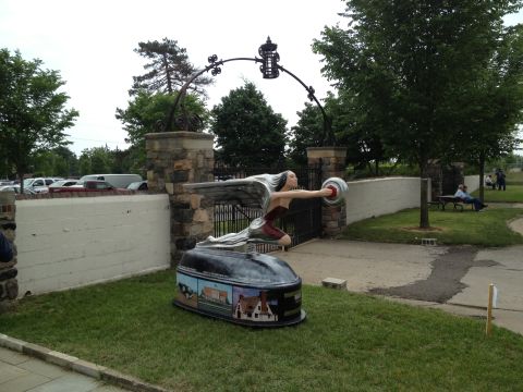 1988 Ford Bronco - A winged woman hood ornament is displayed on a base in front of an arched gateway with parked cars visible behind a distance away.