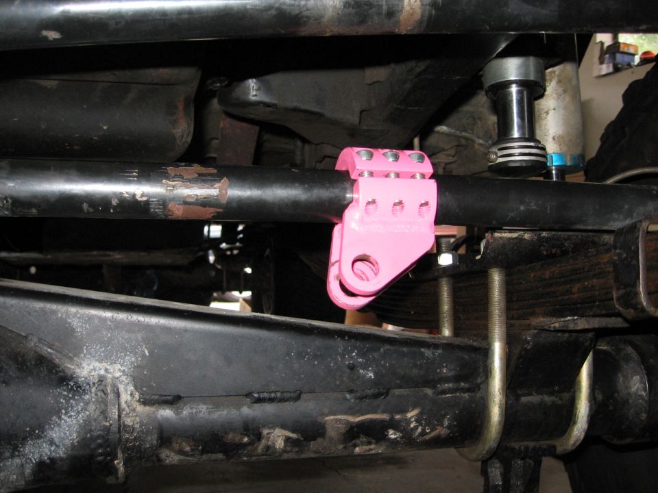 Pink steering stabilizer mount clamped to a black bar on a vehicle's undercarriage. Leaf springs are visible below