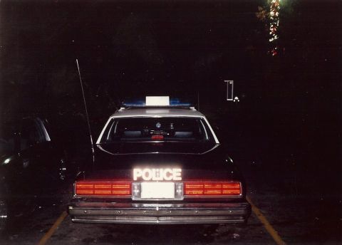 2001 Ford Crown Victoria - A dark police car is parked at night, showing its rear with illuminated "POLICE" text and a light bar on the roof.
