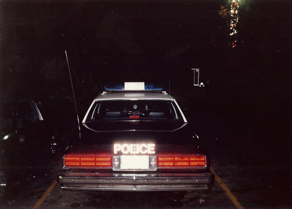 Police car rear view at night, with "POLICE" text illuminated on the trunk. A second vehicle is partially visible to the left