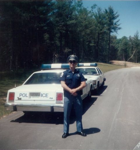 2001 Ford Crown Victoria - Officer poses with two white police cruisers, one showing 'POLICE' on its rear.
