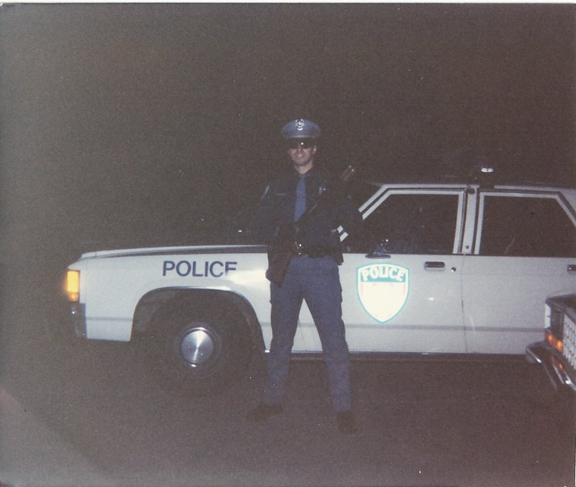 Police officer in uniform standing next to a white police car with 'POLICE' text on the door and a shield emblem