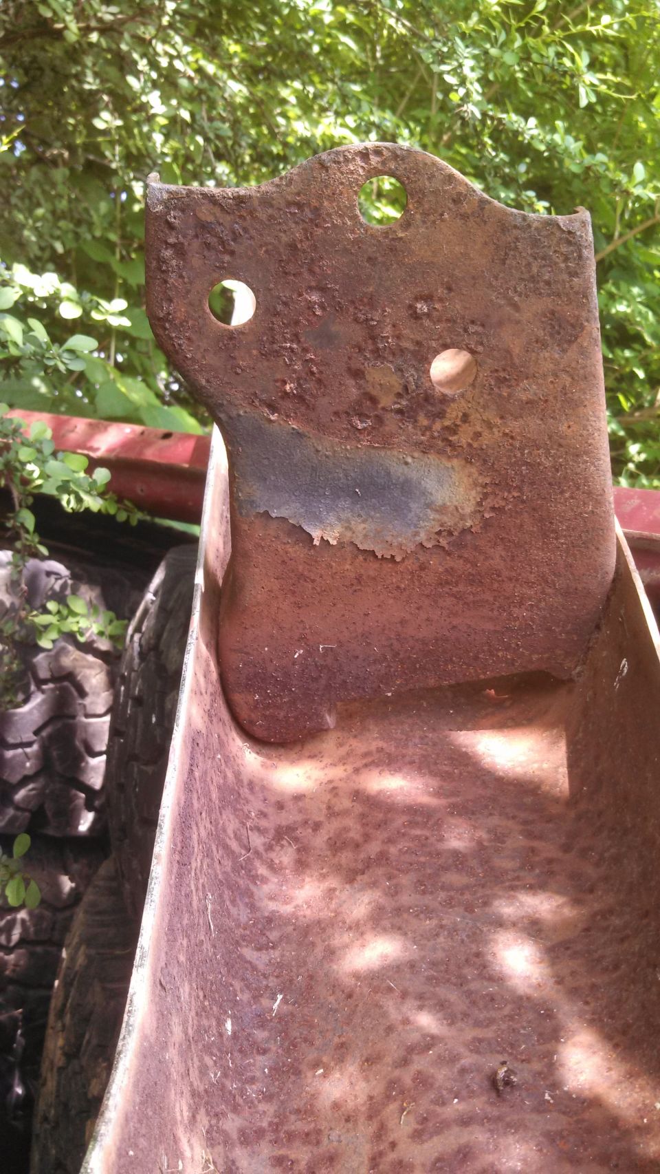 Rusted metal bracket with three holes, one large and two small, against a background of green foliage and a tire
