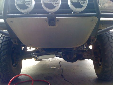 1990 Ford Bronco - Underbody view showing a metal skid plate, steering damper, and three Pro Comp 130 driving lights mounted above.