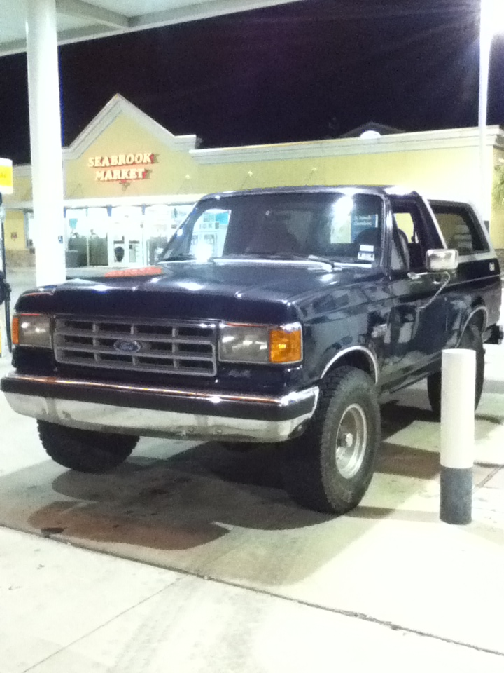 Dark blue Ford Bronco parked at a gas station, front 3/4 view. Visible damage on the front bumper