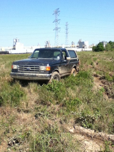 1988 Ford Bronco - A black 1988 Ford Bronco navigates a muddy, grassy terrain with industrial structures in the background.