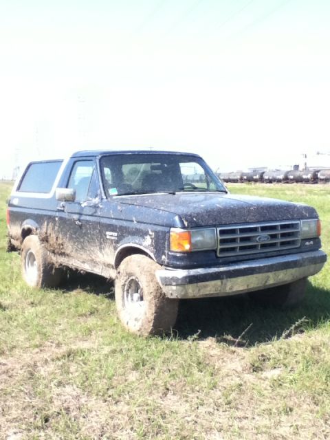 1988 Ford Bronco - A muddy blue Ford Bronco sits in a field under a bright sky, with train cars visible in the distance.