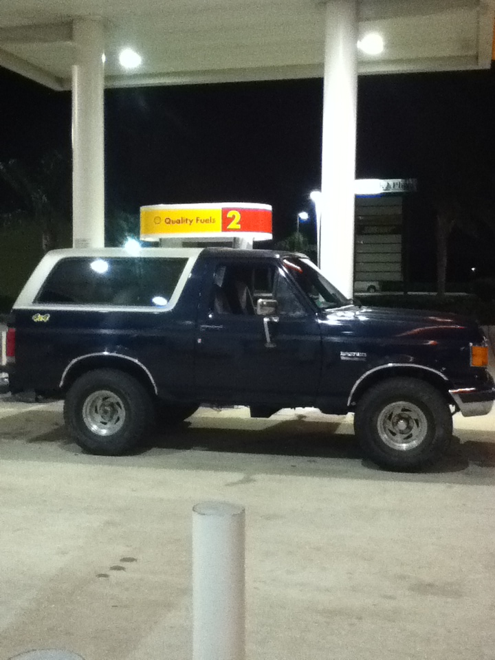 Dark blue Ford Bronco with white top parked at a gas station at night. "Bronco" text visible on fender