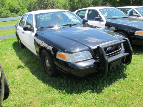 2001 Ford Crown Victoria - A two-tone police car with a push bumper is parked on a grassy lot next to other vehicles.