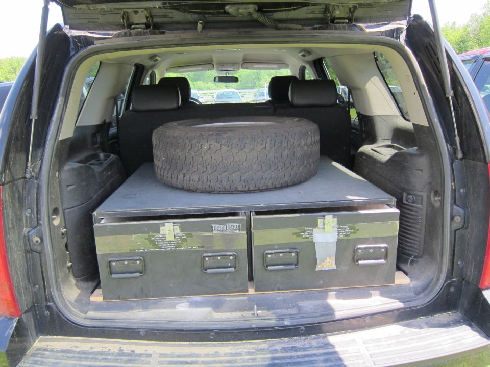 Interior view of black SUV cargo area showing spare tire on top of two green storage boxes
