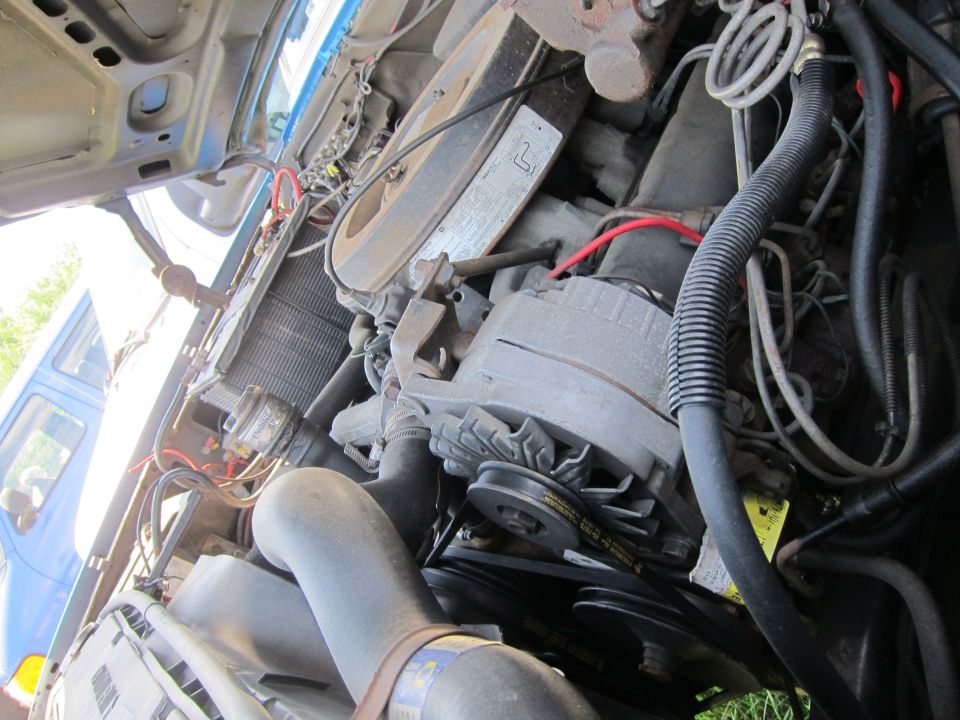 Engine bay with alternator, belts, hoses, and radiator visible. A blue vehicle is partially seen through the grille