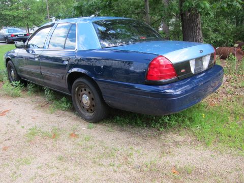 2001 Ford Crown Victoria - A dark blue Ford Crown Victoria with a two-tone roof sits parked on a gravel lot next to overgrown grass.