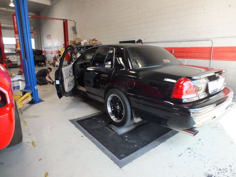 2003 Ford Crown Victoria - black sedan with its door open sits on dyno rollers inside a brick-walled automotive shop with a lift bay.