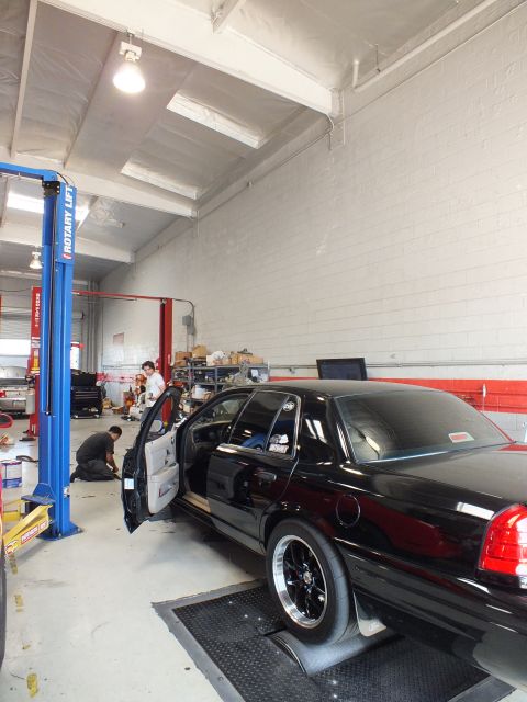 2003 Ford Crown Victoria - black sedan sits with its door open on shop rollers as technicians work nearby in a brightly lit garage.
