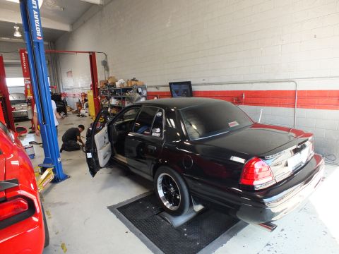 2003 Ford Crown Victoria - black sedan rests on dyno rollers with its door open next to a red car inside a busy automotive shop.