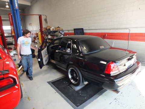 2003 Ford Crown Victoria - man in a white t-shirt stands beside a black sedan with its door open on shop rollers inside a garage.