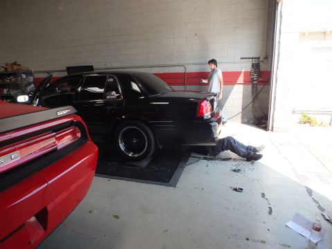 2003 Ford Crown Victoria - black sedan sits on dyno rollers with a person lying beneath the rear as another stands by in the shop.