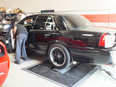 2003 Ford Crown Victoria - technician leans into a black sedan with its door open as it sits on dyno rollers in a brick-walled shop.