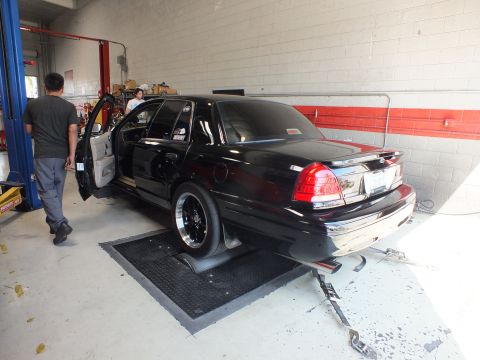 2003 Ford Crown Victoria - rear of a black sedan sits on shop rollers with its door open as a person stands beside it in the garage.