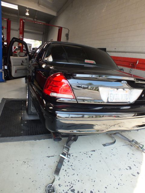 2003 Ford Crown Victoria - rear of a black sedan with an EVIL VIC license plate sits on dyno rollers inside an automotive shop.