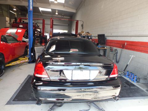 2003 Ford Crown Victoria - rear of a black sedan sits on dyno rollers beside a red car in a shop fitted with two-post lifts.