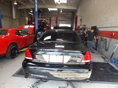 2003 Ford Crown Victoria - rear of a black sedan with an EVIL VIC plate sits on rollers as technicians work beside it in the shop.