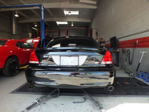 2003 Ford Crown Victoria - rear of a black Ford sedan bearing an EVIL VIC plate and dual exhaust sits on dyno rollers in a shop.