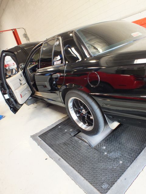 2003 Ford Crown Victoria - side of a black sedan with its door open and a polished drag wheel sits on rollers in an auto shop.