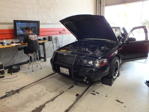 2003 Ford Crown Victoria - black sedan with a front push bar and open hood sits on a dyno facing a technician at a monitor in the shop.