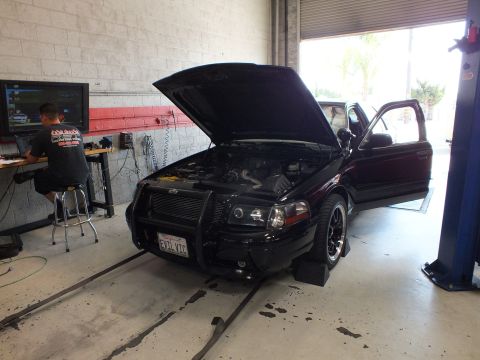 2003 Ford Crown Victoria - black sedan fitted with a push bar and raised hood sits on a dyno as a technician monitors from a desk.