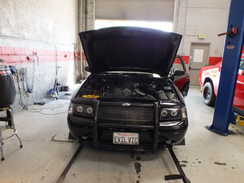 2003 Ford Crown Victoria - front of a black sedan with a push bar, open hood, and EVIL VIC plate sits on a dyno in an auto shop.