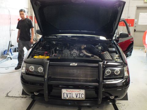 2003 Ford Crown Victoria - black full-size sedan with its hood up and a front brush guard sits on a dyno in a garage, wearing a California "EVIL VIC" Crown Victoria p…