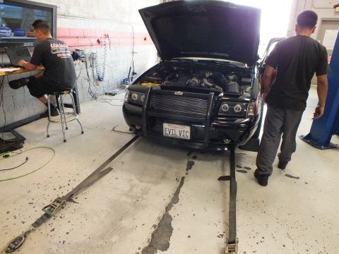 2003 Ford Crown Victoria - black sedan with raised hood and front brush guard sits strapped to a dyno while two men work at a laptop and beside the car in a workshop.