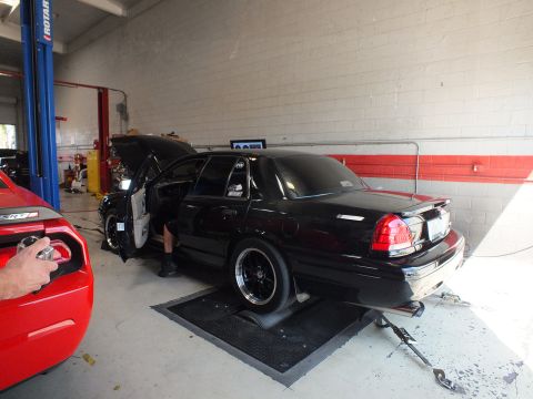 2003 Ford Crown Victoria - Rear three-quarter view of a black sedan with open driver door and wide rear drag wheels parked on a dyno pad inside a workshop.