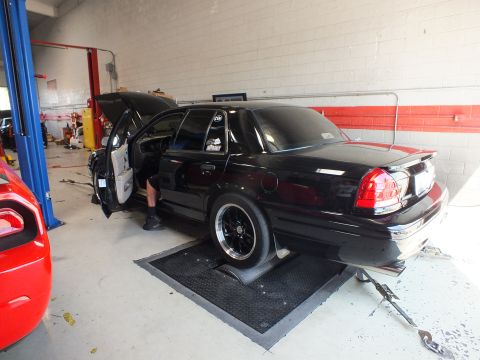 2003 Ford Crown Victoria - black sedan with wide rear drag tires and an open door sits on a dyno pad in a garage with a person crouched at the door.