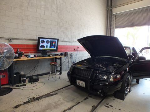 2003 Ford Crown Victoria - black sedan with raised hood and brush guard sits on a dyno facing a wall-mounted dyno readout monitor and a large floor fan in a garage.