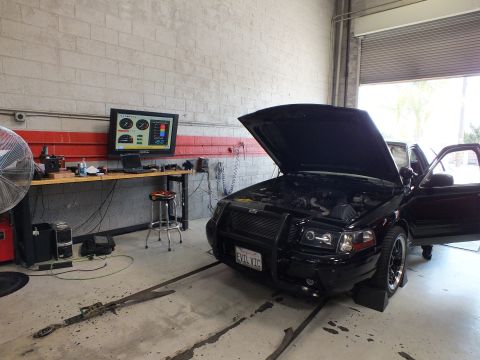 2003 Ford Crown Victoria - black sedan with its hood up and front brush guard sits on a dyno near an open roll-up garage door, with a dyno monitor on the wall behind.