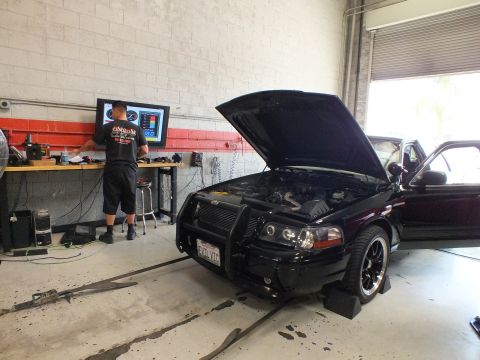 2003 Ford Crown Victoria - black sedan with raised hood and front brush guard sits on a dyno while a man works at a wall monitor in a workshop.